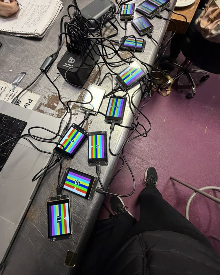 A dozen bare screen modules wired to a 12-port USB charger during burn-in, each showing the firmware's boot test pattern: color bars, its device index, and an up-arrow