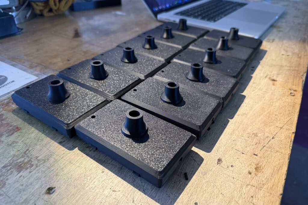 A grid of freshly printed black case base shells on a wooden workbench, each with a stem port glued into its pocket, sockets facing up
