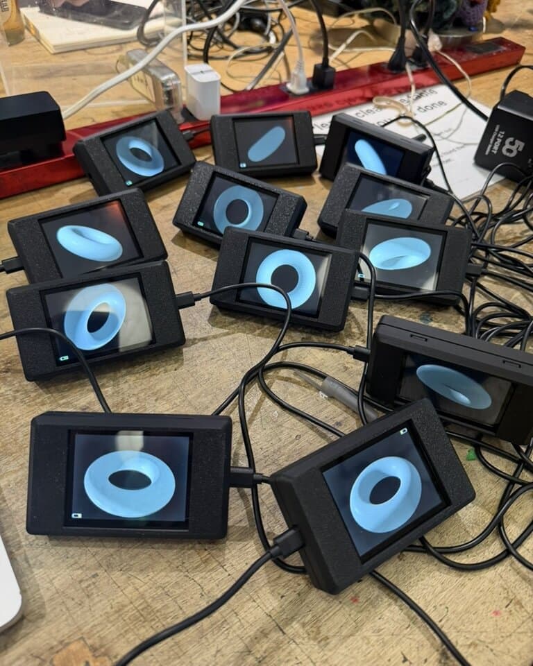 A dozen finished screen modules face-up on the bench, each showing the same blue torus from its own camera's angle